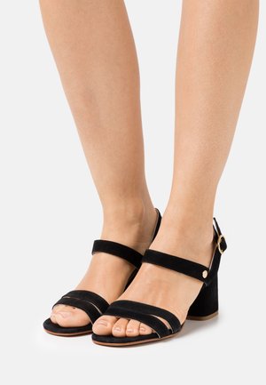 Black suede heeled sandals with two wide straps across the toes, a slim ankle strap, and a chunky heel. Gold-tone hardware detail.