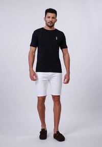 Muž v černém tričku s symbolem esa, bílých šortkách a černých slip-on botách, stojící na světle šedém pozadí.