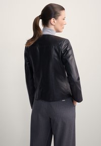 Kobieta z brązowym kucykiem, ubrana w czarną skórzaną kurtkę, szary golf i szare spodnie w prążki, stojąca bokiem na jednolitym tle.