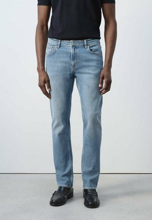 Man wearing light blue slim-fit jeans, black shirt, and black leather loafers standing against a plain light background.