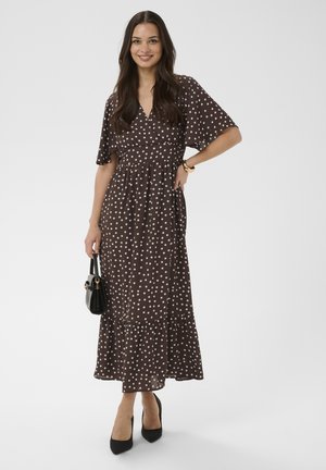 Uśmiechnięta kobieta w brązowej sukience w kropki o długości midi, czarnych szpilkach, trzymająca małą czarną torebkę na jednolitym tle.