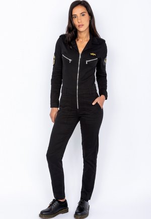 Zwarte jumpsuit met een rits aan de voorkant, lange mouwen en puntige kraag. Beschikt over twee borstzakken en geborduurde patches. Gewaardeerd met zwarte schoenen.