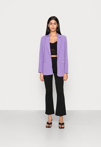 Blazer ajustado roxo com lapelas recortadas, bolsos frontais e um tecido leve. Usado com um top de renda preto cropped e calças pretas largas.