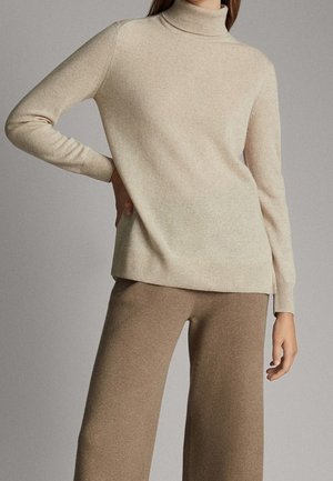 Femme portant un pull beige à col roulé à manches longues et un pantalon marron à jambes larges, debout devant un fond uni.