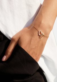 Main avec poignet portant un délicat bracelet en or ornée d'un pendentif cœur ouvert avec de petits diamants, sur fond de vêtements blanc et noir.