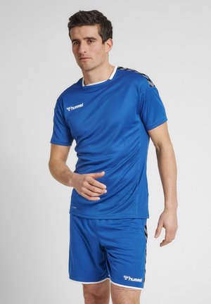 Uomo con indosso abbigliamento sportivo blu con loghi bianchi Hummel sulla maglietta e sui pantaloncini, in piedi davanti a uno sfondo chiaro e uniforme.