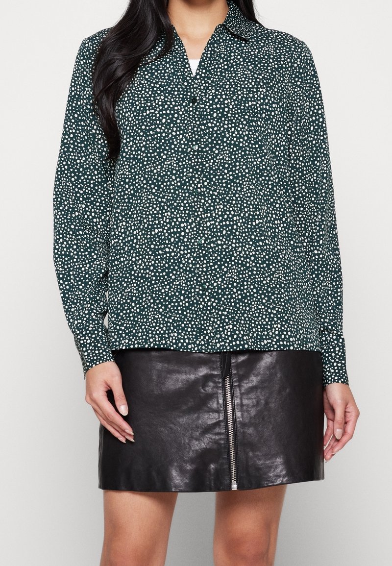 Camicia verde a maniche lunghe con colletto e pois bianchi, abbinata a una mini gonna in pelle nera con zip.