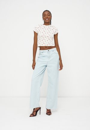 Jeune femme debout portant un jean large bleu clair, un crop top blanc à fleurs et des sandales à talons hauts noires avec des brides, sur fond blanc.