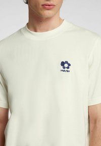Lys beige T-shirt lavet af blødt stof, med rund halsudskæring og korte ærmer, prydet med et lille navy blomsterdiagram på brystet.