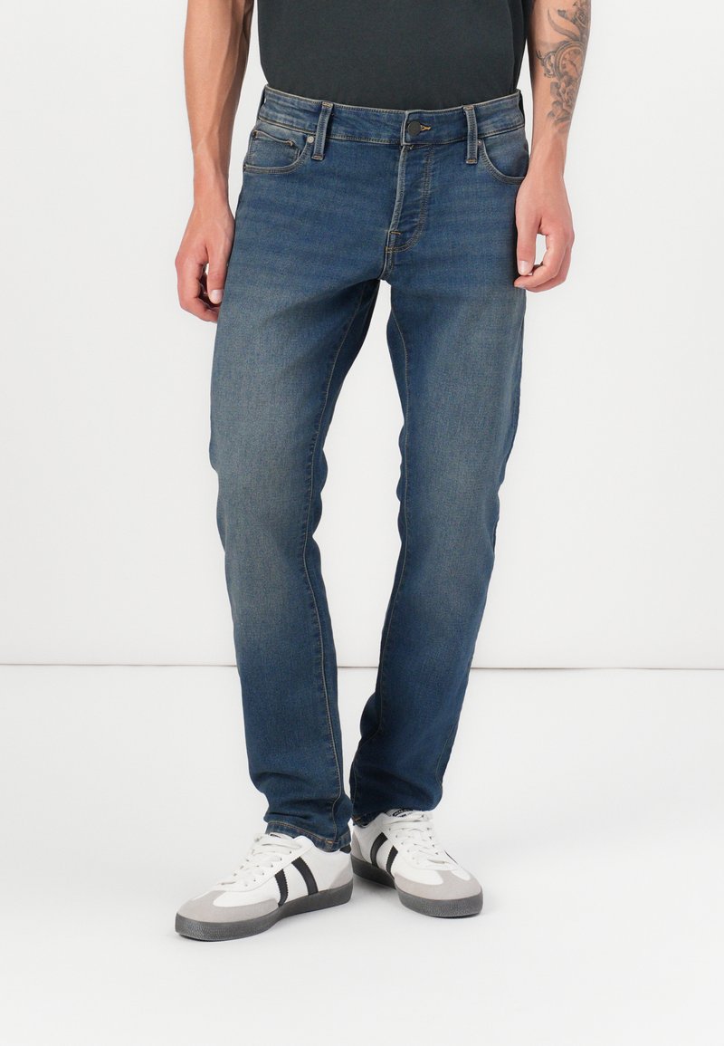 Jeans de denim azul con corte recto, cinco bolsillos y cierre de botón. Combinados con zapatillas grises y blancas con rayas negras.