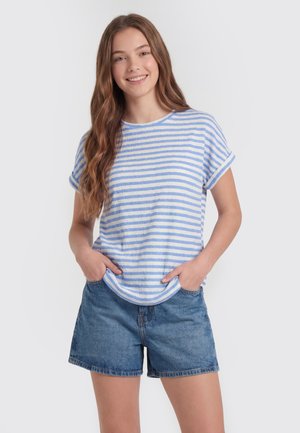 Jonge vrouw met lang bruin haar glimlacht, draagt een blauw-wit gestreept T-shirt met korte mouwen en denimshorts, handen in de zakken.