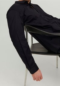 Camicia a maniche lunghe, con bottoni, in cotone nero unito, con colletto classico e una texture liscia, indossata mentre si è seduti su una sedia.