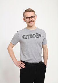 Šedé bavlněné tričko s potiskem "CITROËN" v černé barvě, spárované s černými kalhotami a černým pásem. Jednoduchý, neformální design bez vzorů.