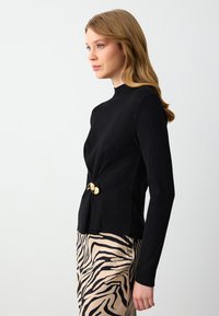 Zwarte ribgebreide trui met hoge hals, voorzien van een gouden knoopaccent ter hoogte van de taille. Gecombineerd met een beige rok met zwarte zebrapatronen.
