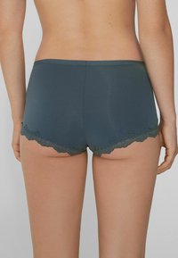 Pantalones cortos hipster en verde azulado oscuro con un borde de encaje ondulado, tejido suave y un diseño ajustado, mostrando la vista trasera.