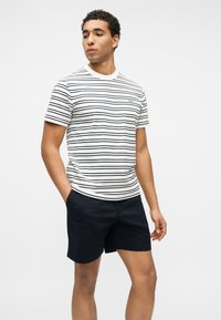 Άνδρας που φοράει λευκό και μαύρο ριγέ κοντομάνικο πουκάμισο και μαύρο σορτς, στέκεται με το ένα χέρι στη τσέπη μπροστά σε μονόχρωμο φόντο.