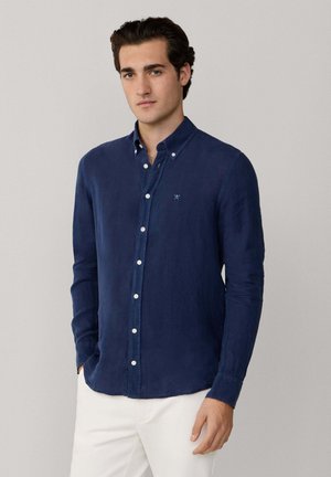 Jonge man met donker haar, gekleed in een donkerblauw overhemd met knopen en een witte broek, staand tegen een effen lichtgrijze achtergrond.