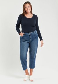 Granatowa bluzka z długim rękawem w połączeniu z jasnoniebieskimi spodniami typu high-waist o luźnym kroju i skróconej długości, dopełniona białymi klapkami na odkrytej stopie.