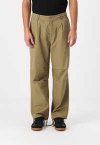 Lightweight, khaki-colored trousers with a relaxed fit, featuring a buttoned waistband and subtle front pleats. Worn with black sneakers.