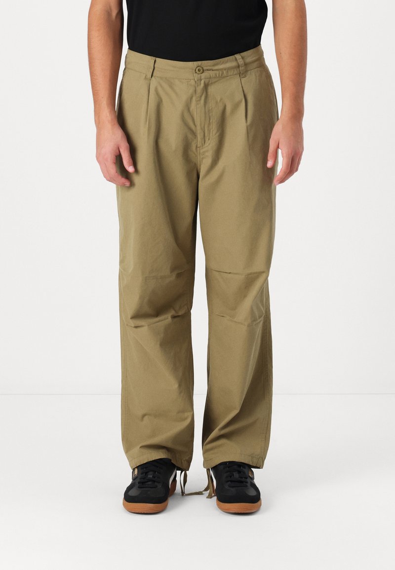 Lightweight, khaki-colored trousers with a relaxed fit, featuring a buttoned waistband and subtle front pleats. Worn with black sneakers.