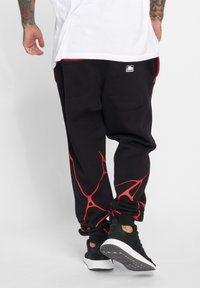 Black sweatpants with red abstract design, featuring two back pockets and elastic cuffs, paired with black athletic shoes.