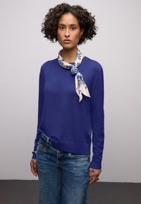 Pull violet à manches longues, avec poignets et ourlet côtelés. Associé à un jean en denim bleu. Une écharpe crème à motif floral bleu est nouée au cou.