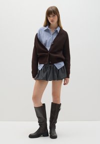 Cárdigan marrón de punto, camisa de botones rayas azules, falda acampanada gris y botas de cuero marrón hasta la rodilla, con detalle de cremallera.