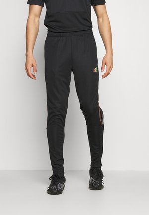 Mężczyzna w czarnych spodniach sportowych Adidas z kolorowym logo na prawej udzie oraz czarnych butach sportowych z białymi wzorami.