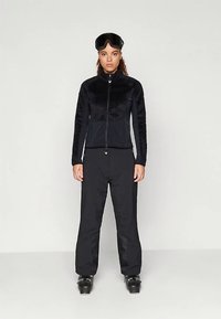 Naine kannab musta suusajakki, musti suusapükse, musti suusasaapaid ja suusaglasse, mis on tal otsaesisel, seismas ühetaolise tausta ees.