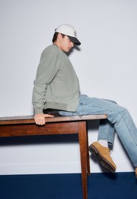 Lysegrøn bomberjakke, cowboybukser, brune sko med teksturerede såler og en hvid kasket med mørk skygge, sidder på et træbord.