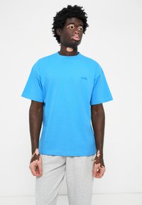 Ung mand med vitiligo iført en blå t-shirt og lysegrå sweatpants, stående mod en simpel hvid baggrund og kigger til siden.