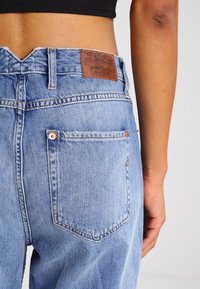 Jeans de mezclilla azul claro con cintura alta, que presentan un parche de cuero en la parte posterior, dos bolsillos traseros y costuras en contraste.