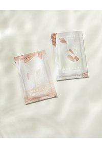 Deux sachets de masque facial sur une surface claire. L'un est rose avec des accents floraux ; l'autre est blanc avec des graphiques subtils. Les deux affichent une marque.