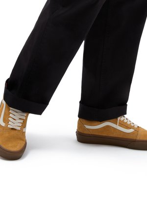 Zapatillas de ante de color marrón con franjas laterales blancas, suelas de goma marrón y cordones color crema; combinadas con pantalones negros con puños.
