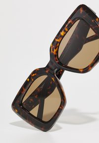 Occhiali da sole oversized con montatura in tartaruga, lenti marroni e forma angolare spessa. Superficie testurizzata con finitura lucida e dettagli del marchio sottili.