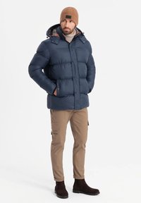 Giacca imbottita blu scuro con cappuccio e fodera a quadri, abbinata a pantaloni cargo marrone chiaro e stivali scamosciati marrone scuro.