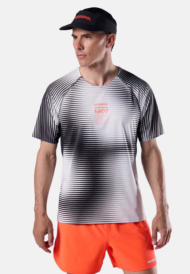 Mann trägt ein schwarzes und weißes, gestreiftes Rossignol-T-Shirt mit Verlauf, leuchtend orangefarbene Shorts und ein schwarzes Rossignol-Visier, steht links und ist vor einem einfarbigen Hintergrund abgebildet.