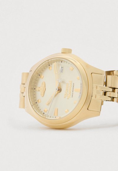 Zelta toņa Vivienne Westwood rokas pulkstenis ar metāla siksniņu, dimanta formas stundu marķieriem, datuma logu un Šveices mehānismu uz baltas virsmas.
