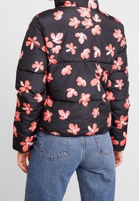Svart pufferjacka med blommönster i rosa och vita detaljer. Materialet är glansigt, med vadderade sektioner som tillför textur.