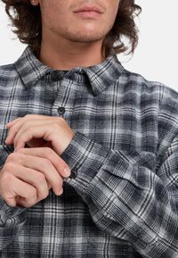 Czarna i biała flanelowa koszula w kratę z kołnierzykiem, zapinanymi na guziki mankietami i teksturowaną tkaniną. Charakteryzuje się wyraźnym kratkowanym wzorem i czarnymi guzikami.