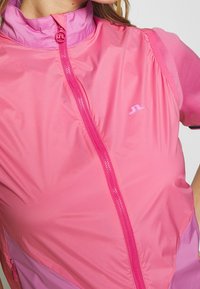 Růžová lehká bunda s vysokým límcem, plným předním zipem a barevnými bloky. Obsahuje malé logo na hrudi.