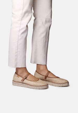 Nohy obuté v béžových tkaných espadrillách s hnedými remienkami a prackami, v kombinácii s krémovými krátkymi nohavicami.