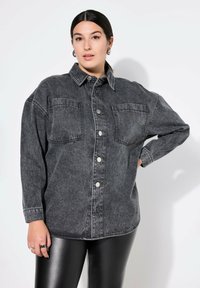 Camisa de mezclilla negra con botón en la parte delantera, dos bolsillos en el pecho, corte relajado y mangas largas. Textura suave con un mínimo de desgaste.