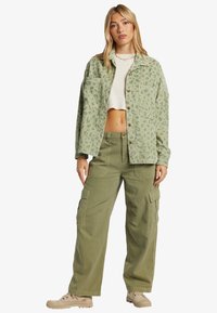 Chaqueta verde de corduroy con estampado de animales, cierre de botones y dos bolsillos frontales, combinada con pantalones cargo oliva que cuentan con bolsillos laterales.