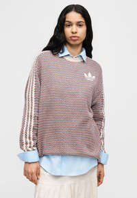 Πλεκτό πουλόβερ σε πορτοκαλί, μπλε και λευκό με στρογγυλή λαιμόκοψη, διακοσμημένο με λευκό λογότυπο και ριγέ μανίκια. Φορεμένο πάνω από ένα ανοιχτό μπλε πουκάμισο.