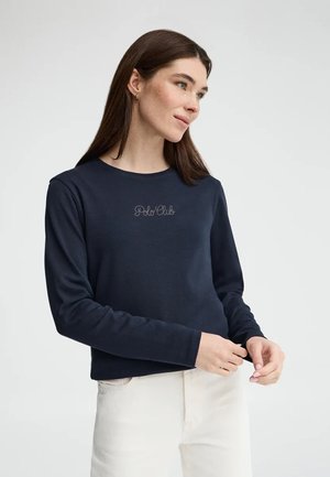 Frau mit langen braunen Haaren, die einen dunkelblauen "Polo Club"-Pullover und eine weiße Hose trägt, stehend vor einem einfarbigen hellen Hintergrund.