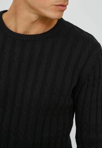 Pull en tricot noir avec un motif en câble texturé, un col rond côtelé et des manches longues, mettant en avant une coupe ajustée.