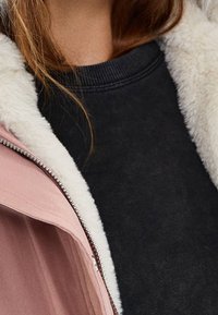 Lyserød jakke med et shearling-kraveskilt, åbent for at vise en sort sweatshirt under. Bløde teksturer og kontrasterende farver er synlige.