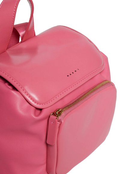 Sac à dos en cuir rose avec une texture lisse, fermeture éclair dorée, poche avant et nom de marque embossé. Dispose de bretelles doubles pour le porter.