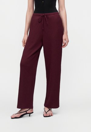 Persoon die losse, gestructureerde donkerrode broek met trekkoord in de taille en zwarte, sandalen met bandjes en lage hak draagt op een lichte achtergrond.